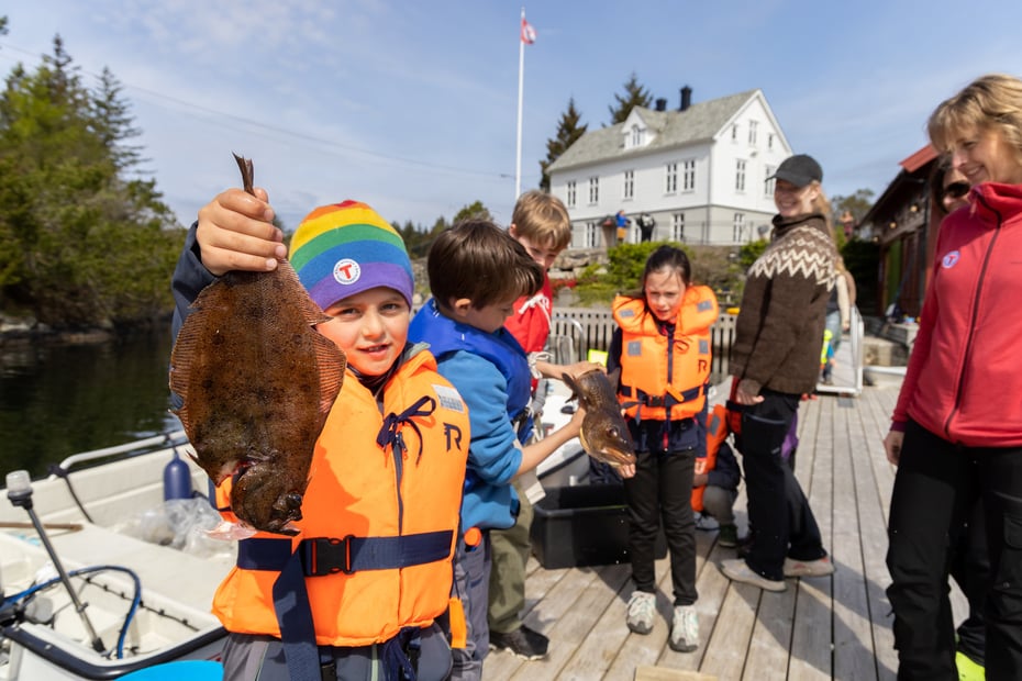 Gutt holder opp en flyndre foran en hytte og flere andre barn og voksne bak på bryggen.
