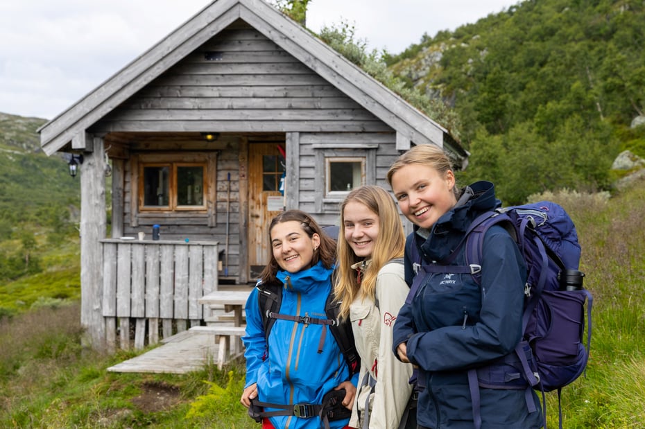Tre jenter i turklær og ryggsekk står foran en gammel, fin hytte og smiler.