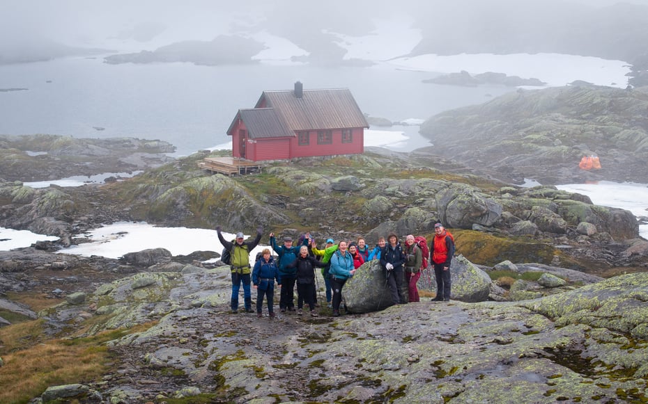 En glad og jublende turgjeng står foran en rødmalt hytte i tåkekledd fjellområde med noen snøflekker rundt hytten.
