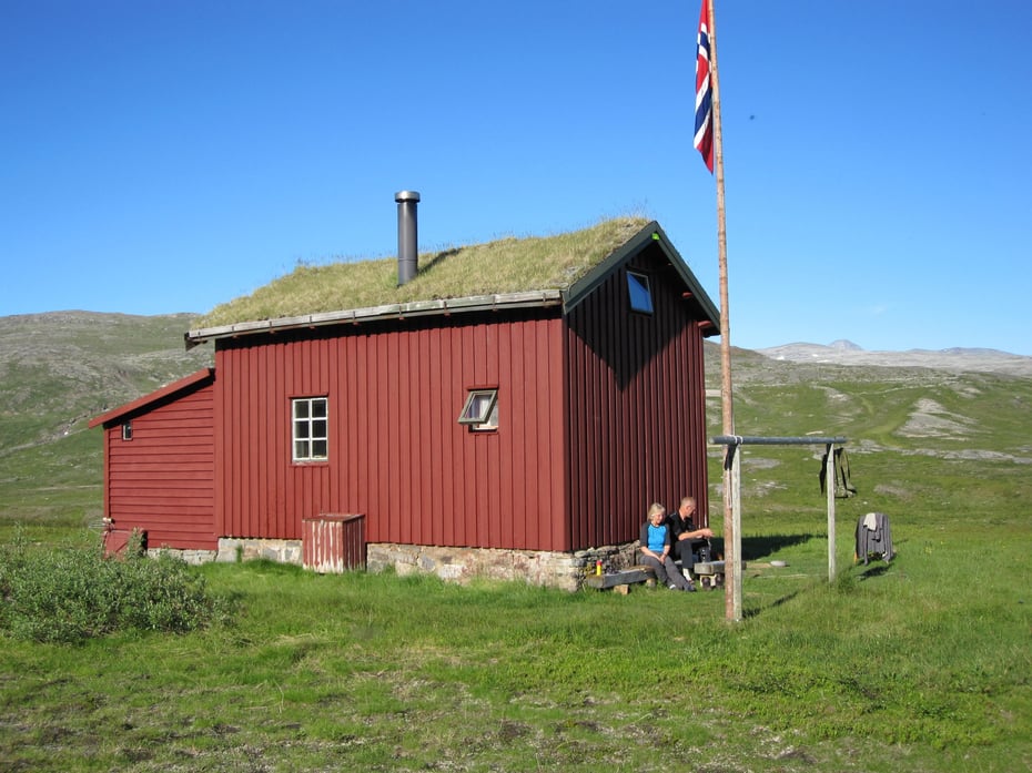 Ei rødmalt hytte med torvtak. Det er ei flaggstang med norsk flagg foran hytta. To turkledte mennesker sitter langs veggen. Det er sommer og grønt.