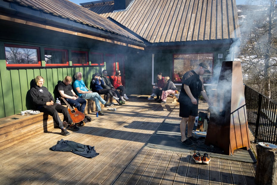 Folk slapper av på terrasse utenfor hytte i solen mens en mann griller.