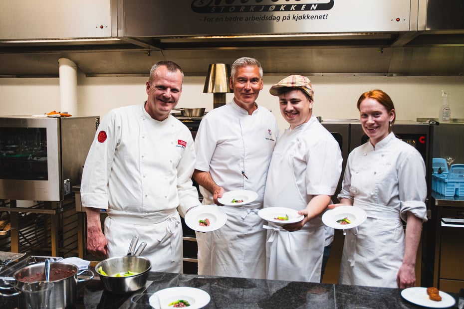 Fire kokker fra de betjente anleggene i Stavanger Turistforening poserer foran kamera på Norrønna storkjøkken i Stavanger. Foran seg holder de danderte retter. 