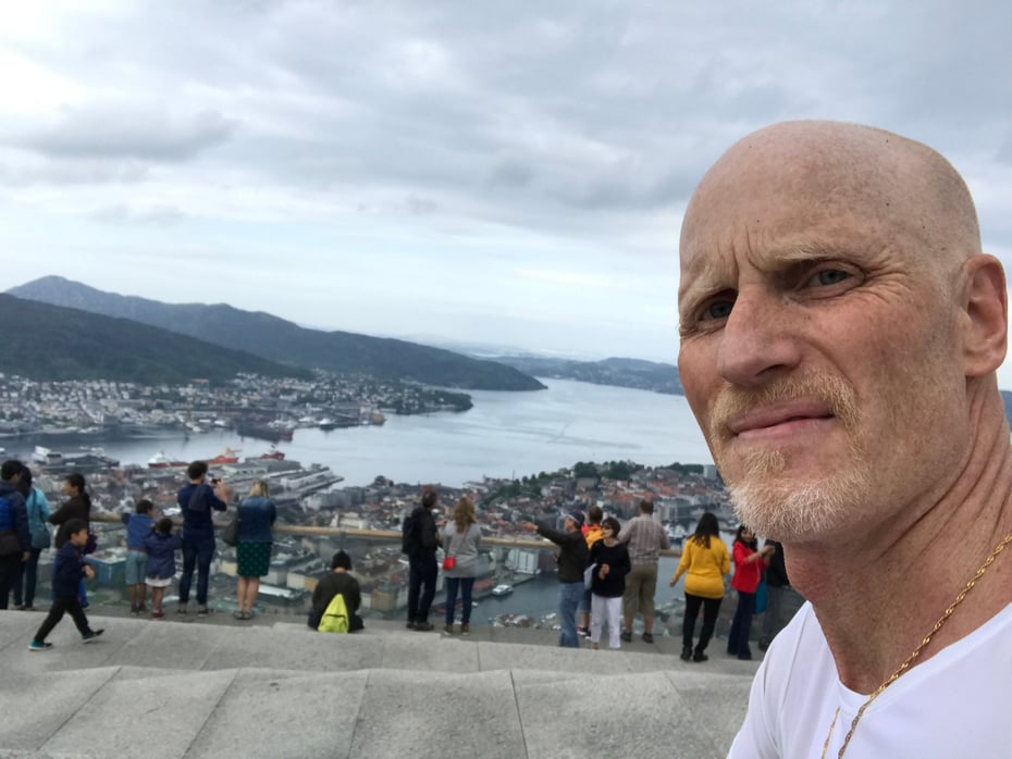 Man i hvis t-skjorte ser inn i kamera på toppen av Fløyen med Bergen bak.