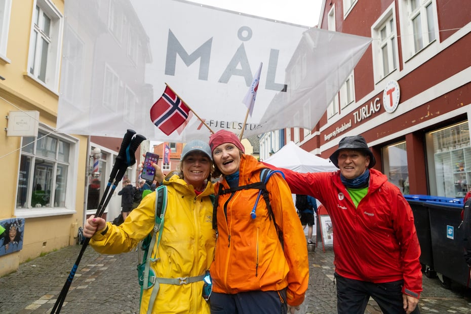 To glade turgåere i mål med en frivillig som holder opp norsk flagg bak dem.