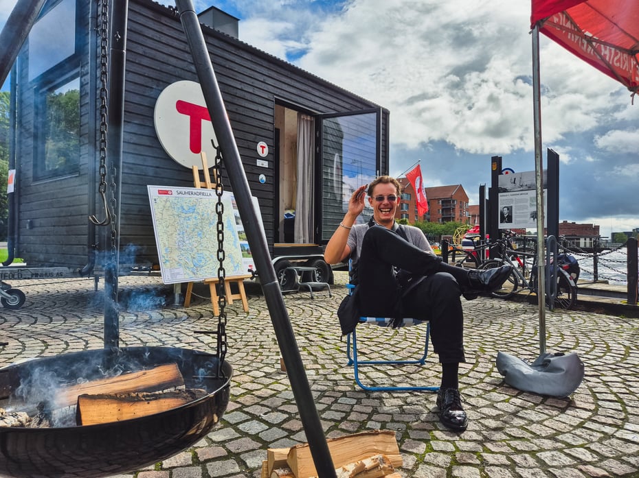 En smilende mann sitter foran en hytte i byen med en bålpanne ved siden av