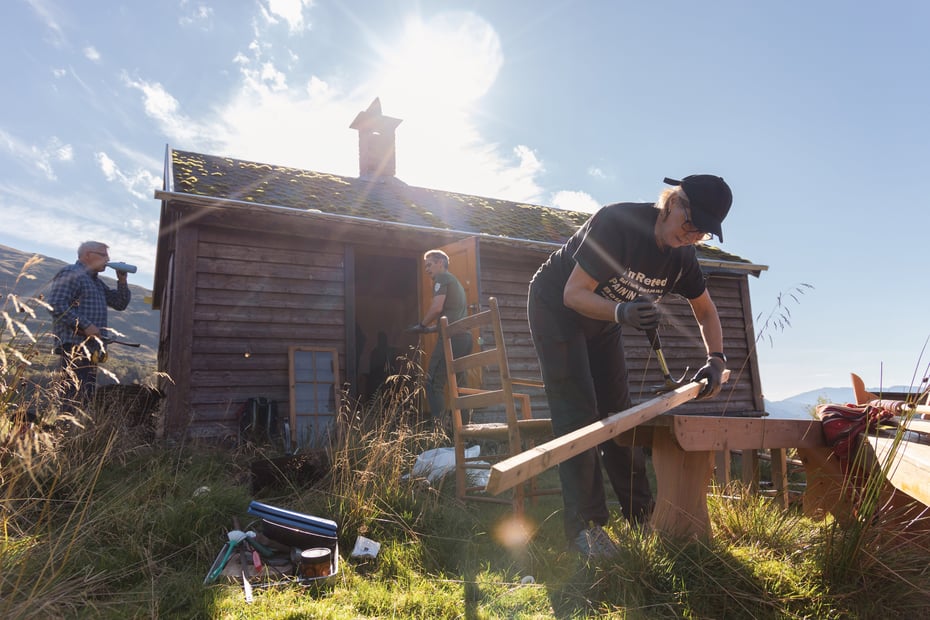 En dame med hammer jobber dugnad foran en hytte i høstsolen.