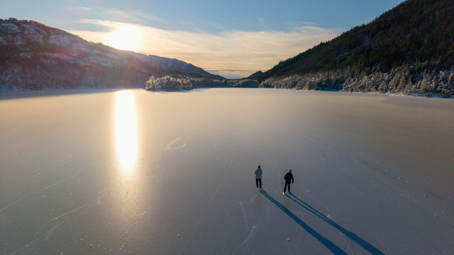 To personer på skøyter på islagt vann i nydelig vinterlandskap.