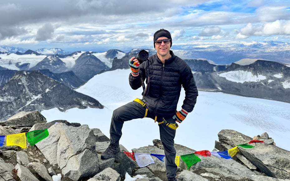 Mann med kamera på Galdhøpiggen med utsikt over mange fjell.