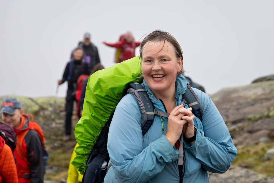 Kvinne med sekk smiler mot kameraet på fjelltur med mennesker bak. 