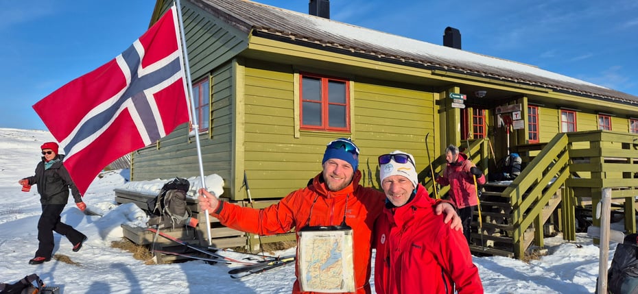 To menn med norsk flagg foran hytte om vinteren og ski i bakgrunnen.