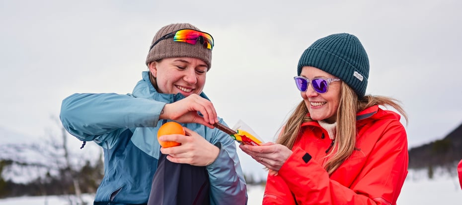 To kvinner sitter ute i vinterlandskap. De smiler og en holder en appelsin menst hun får sjokolade av den andre kvinnen. 