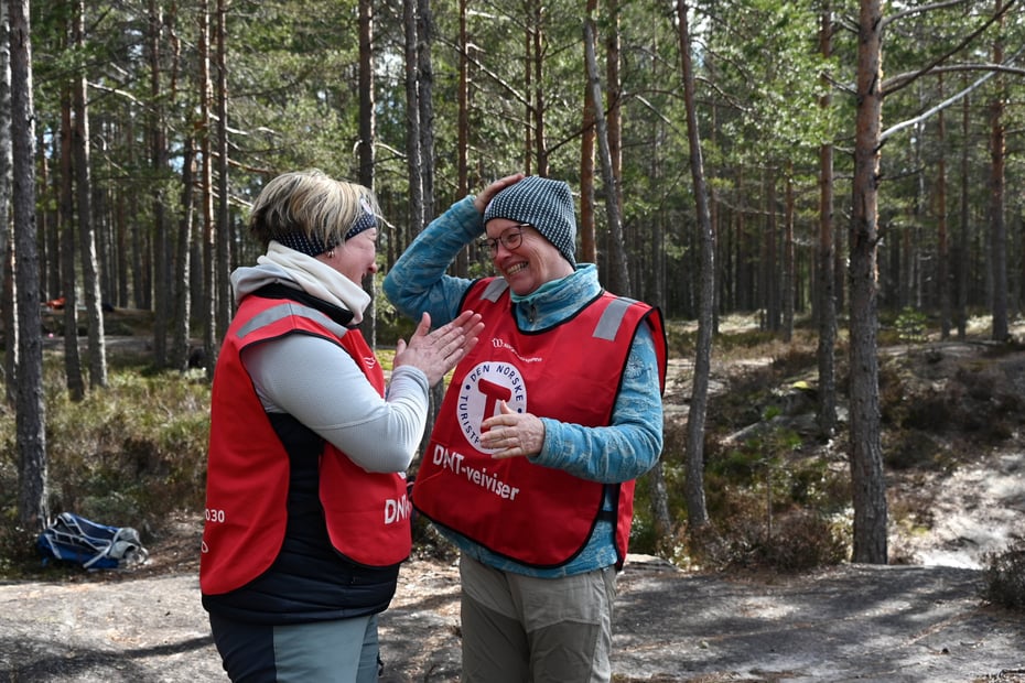to damer med rød dnt-vest står i skogen og smiler til hverandre