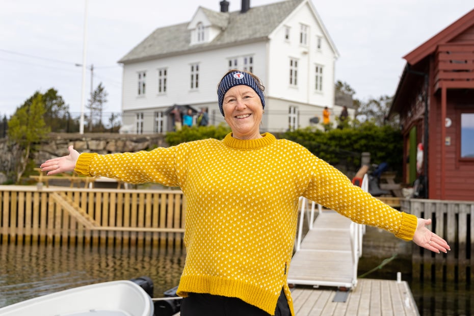 Dame i gul ullgenser slår armene ut foran hvit hytte ved sjøen.