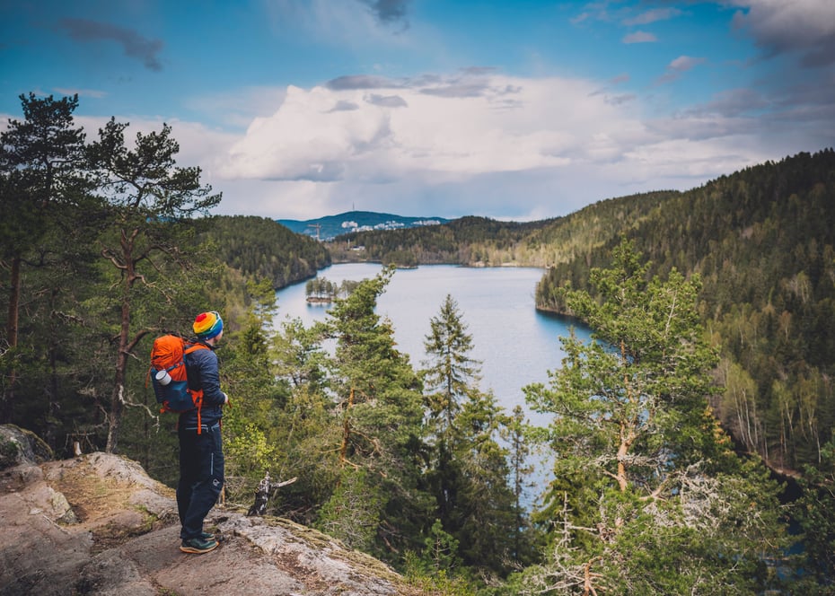 mann med sekk og regnbuelue som står på en høyde og ser utover et vann og skog