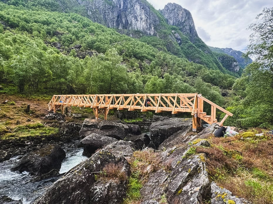 En trebro over en elv i en dal. Skog og høye fjell i bakgrunnen.