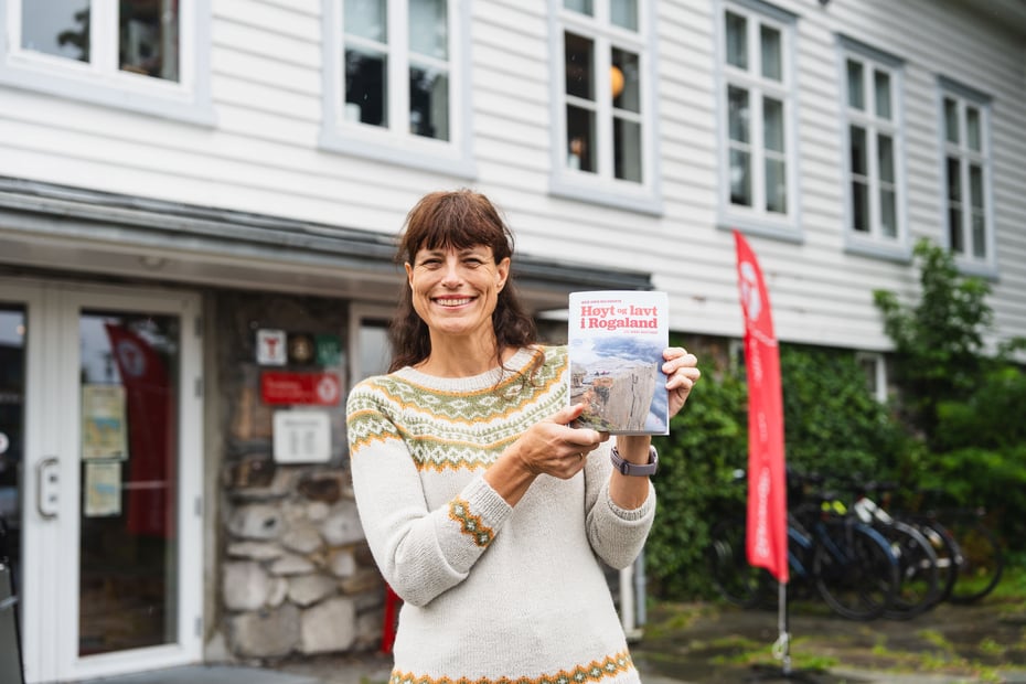 Dame i ullgenser holder bok i hånden utenfor hvitt hus