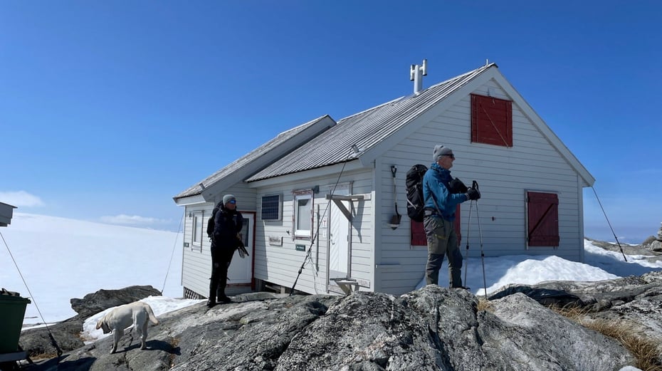 Lysegrå hytte ved isbre og folk foran på ski.
