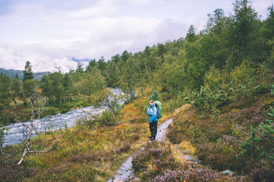 En person står på en sti. Det er en elv til venstre og det er mye vann i elven. Det har regnet og hun er godt kledd med hetta oppe og sekk på ryggen. 