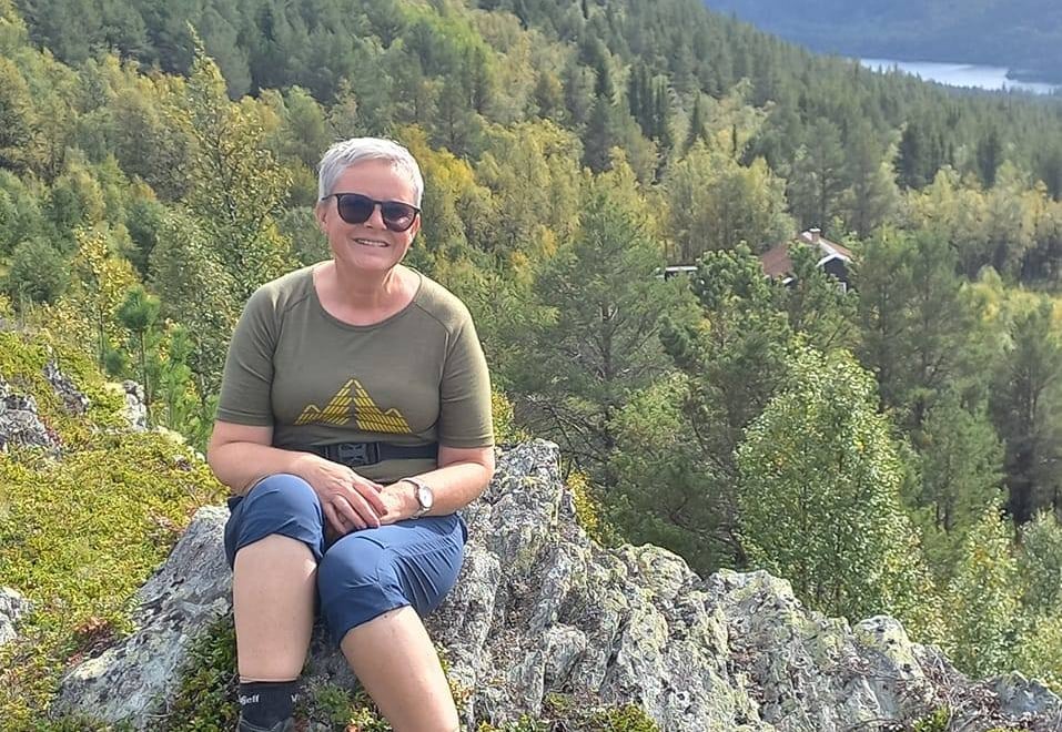 Smilende kvinne i turbekledning som sitter på en fjellknaus med utsikt i bakgrunnen. Foto.