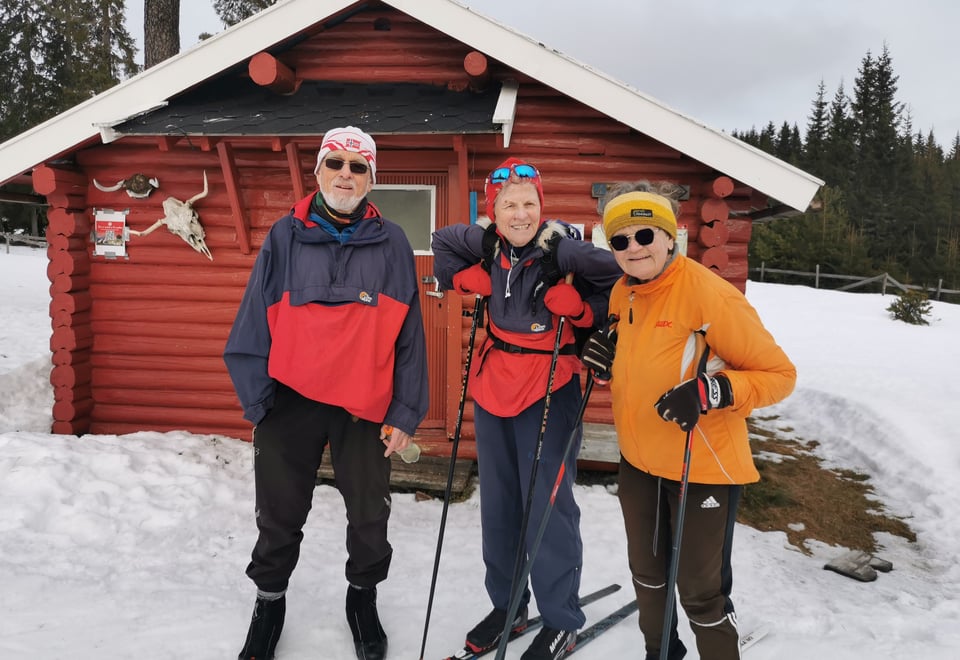 Tre blide eldre mennesker utenfor rød koie.