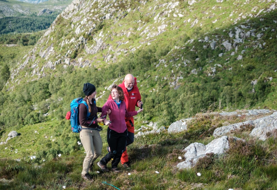 Tre personer går i fjellterreng. Damen i midten har en gul bøyle som hun holder i og blir støttet av to ledsagere. Alle personene smiler og ler.