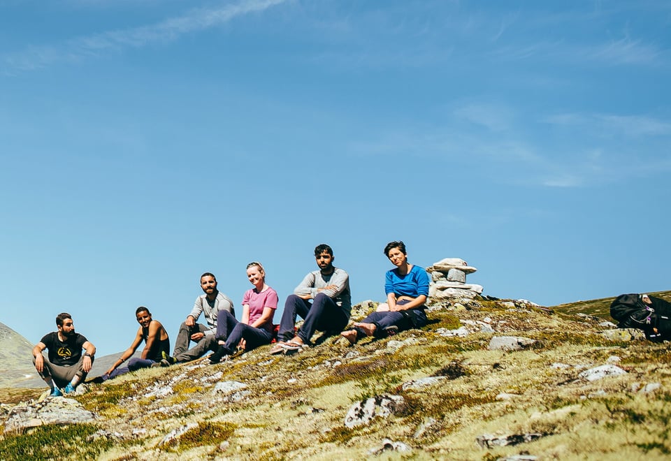 En gruppe mennesker sitter på en fjellrygg, men høye fjell i bakgrunnen.