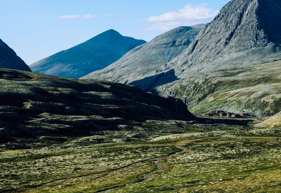Et fjellandskap med steinete og kuperte fjell som strekker seg mot horisonten. I bunnen av dalen kan man se noen små bygninger som står samlet, omgitt av grønt landskap og lyng. Fjellene i bakgrunnen gir et majestetisk og dramatisk inntrykk, mens noen områder fortsatt har små flekker av snø.