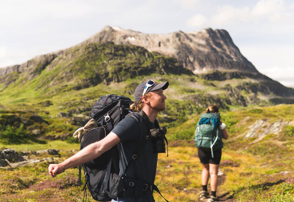 To personer ute på tur, begge med stor tursekk på ryggen. Stort fjell i mellomgrunnen