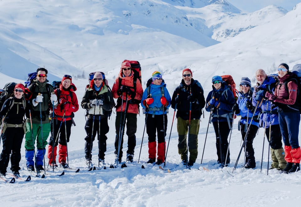 A group of smiling hikers equipped with backpacks and ski poles, dressed in winter gear, stand together on a snow-covered mountain landscape, ready for a winter adventure under clear skies