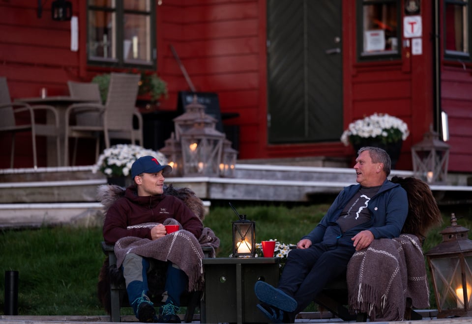 To menn sitter i hver sin stol utendørs i kveldssola og snakker sammen. Lykter med stearinlys er tent.