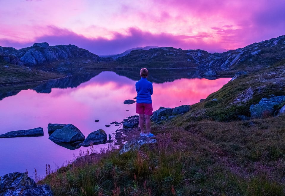 Menneske som står på et berg ved et vann mens solnedgangen lager himmelen rosa og lilla.
