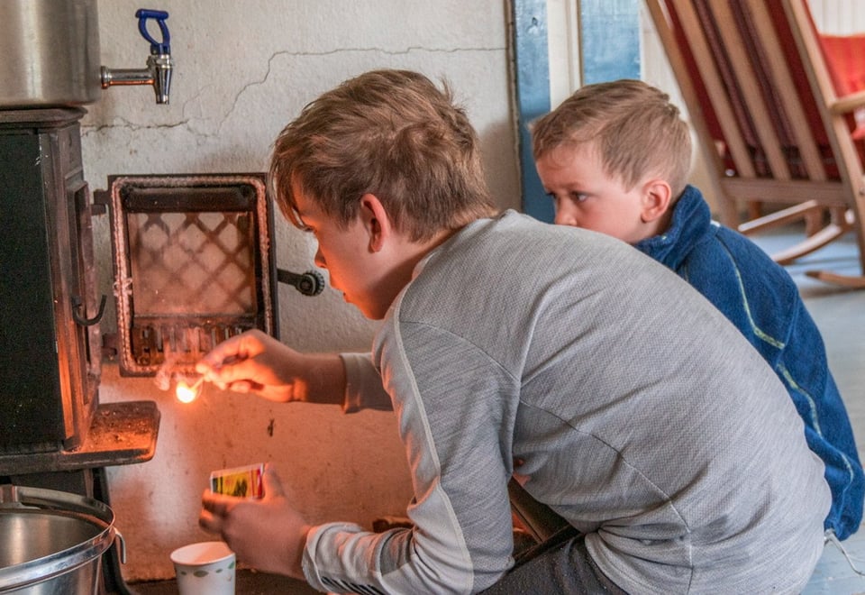 To gutter som fyrer i en vedovn. Den ene gutten har en fyrstikk i hånden.