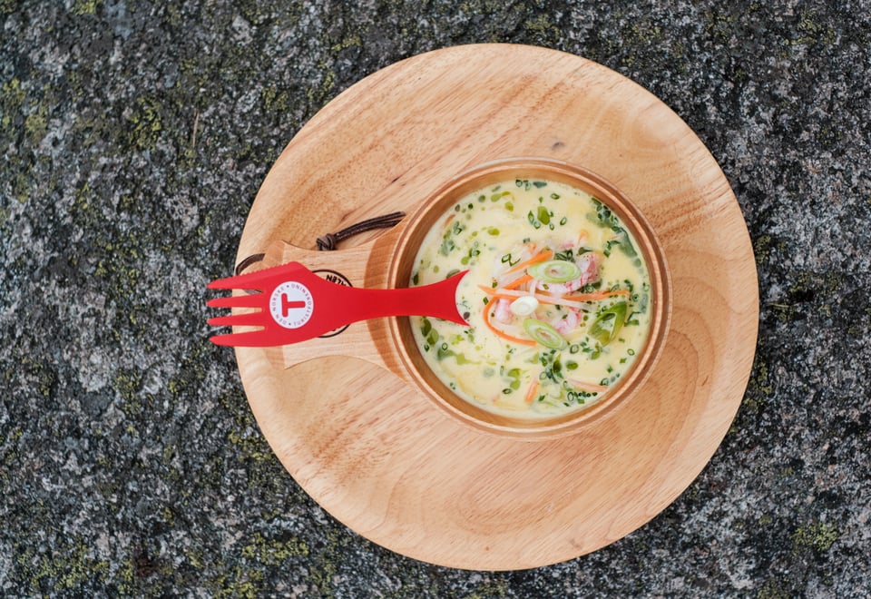 En tretallerken med en trekopp som inneholder fiskesuppe. Oppi suppa er det en rød skei med en T-logo på.