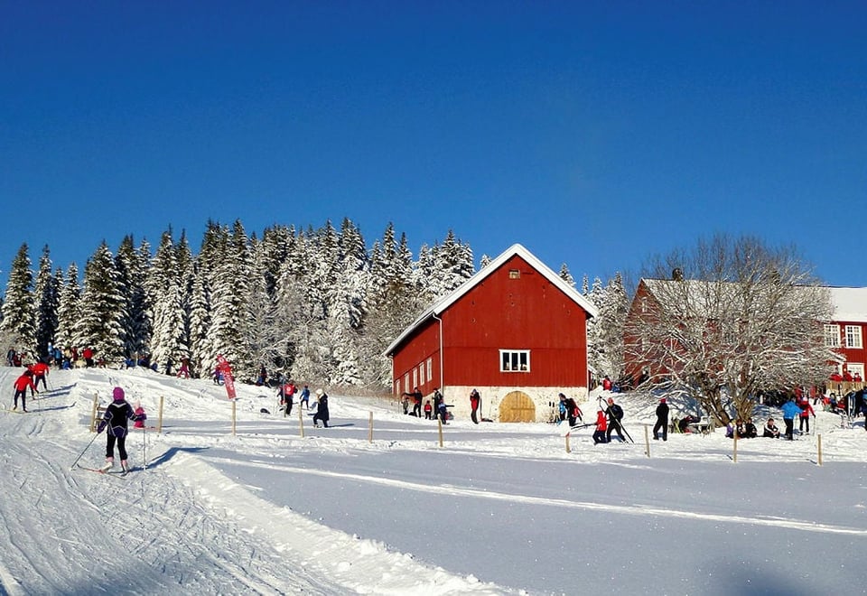 En stor rød hytte og en rød låve i utkanten av en skog, vinter og masse snø på trærne. Mennesker går i oppkjørte skispor foran hytta.