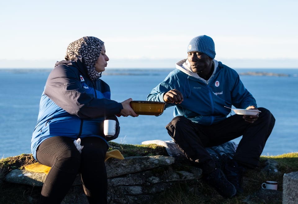 To personer sitter på toppen av et fjell og deler en matpakke. Begge har på seg blå allværsjakker hvor det står dugnad på.
