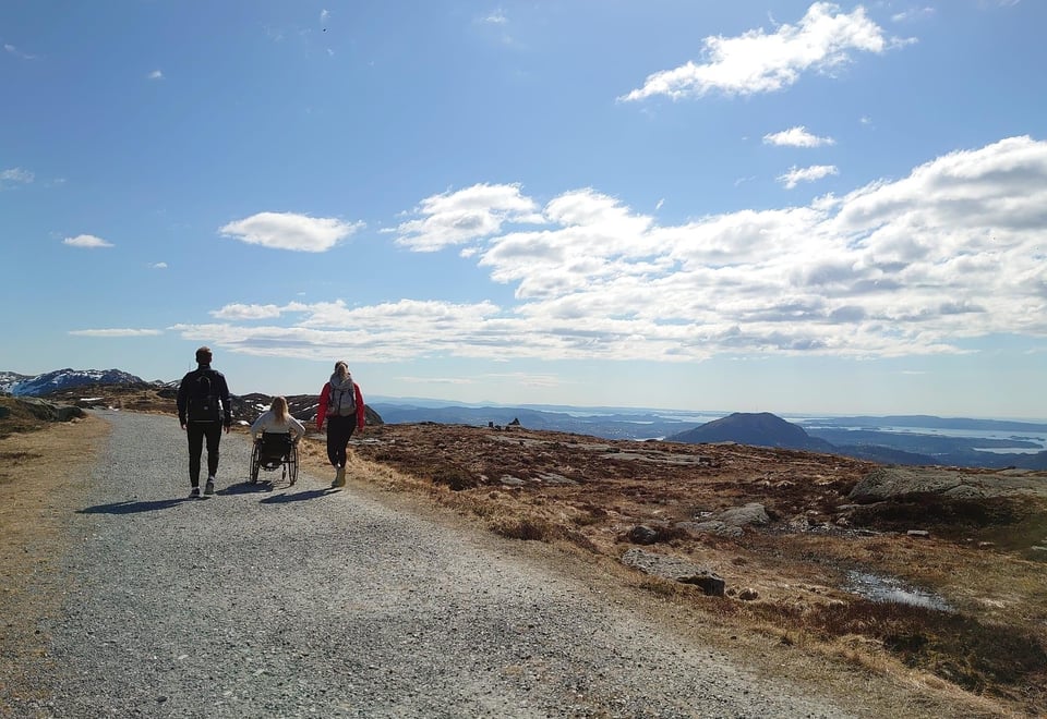 To gående og en trillende på tur på grusvei, med blå himmel og fjell i bakgrunnen