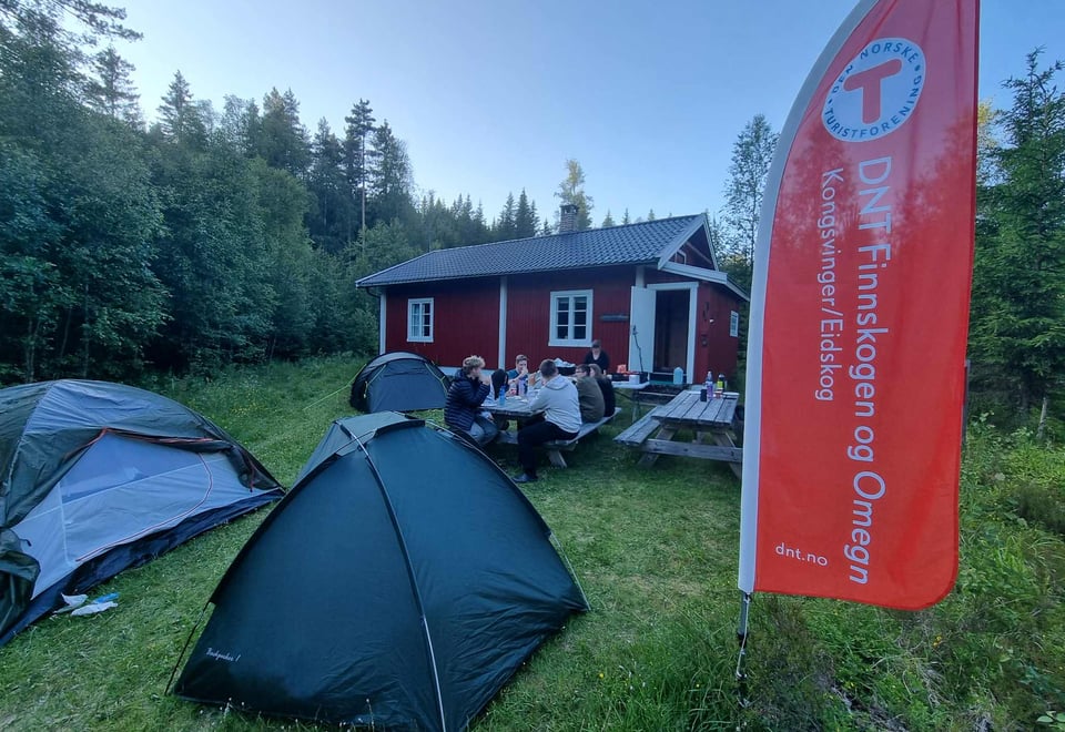 Telt, ungdommer som spiser foran DNT-hytte med beach flag i forkandt