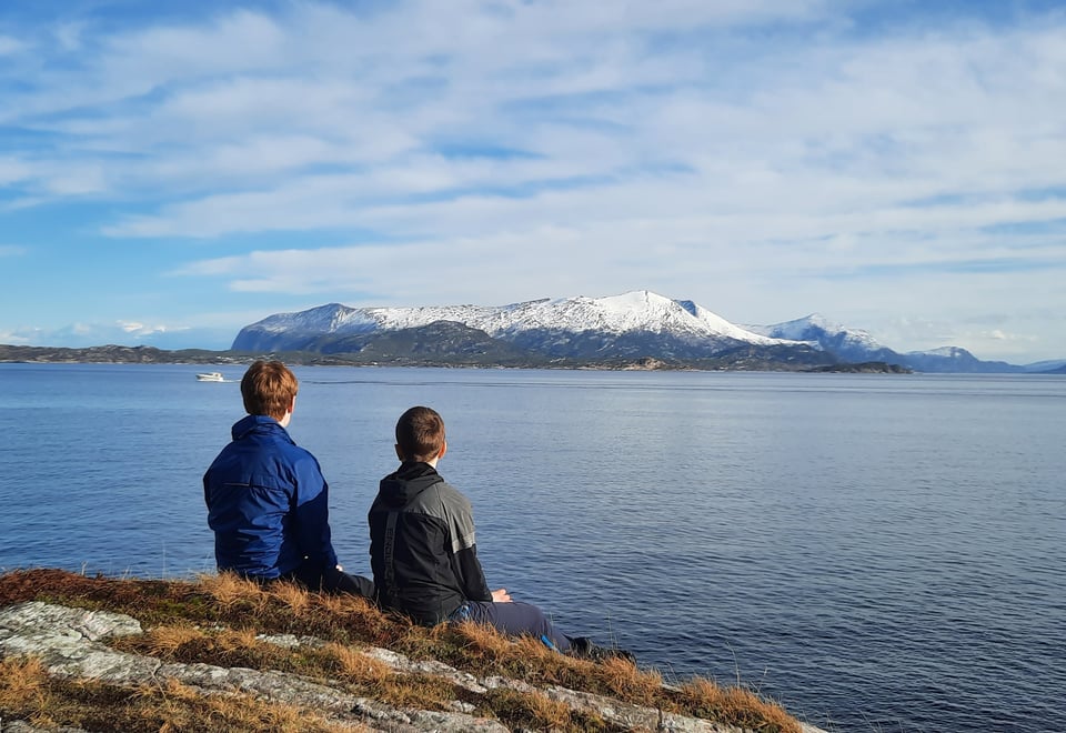 To personer sitter på berget på Sandvikstien i Kristiansund med ryggen til. De har utsikt mot Tustnafjella