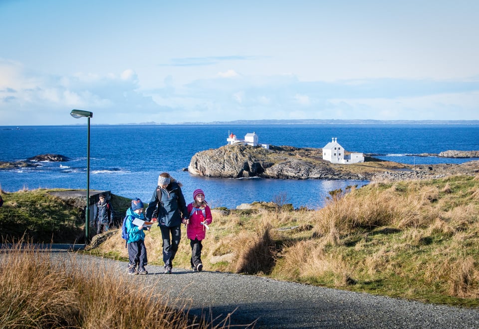 En kvinne med to barn går oppover en grusvei. De holder hender. Bak dem skimtes et fyr og blått hav.