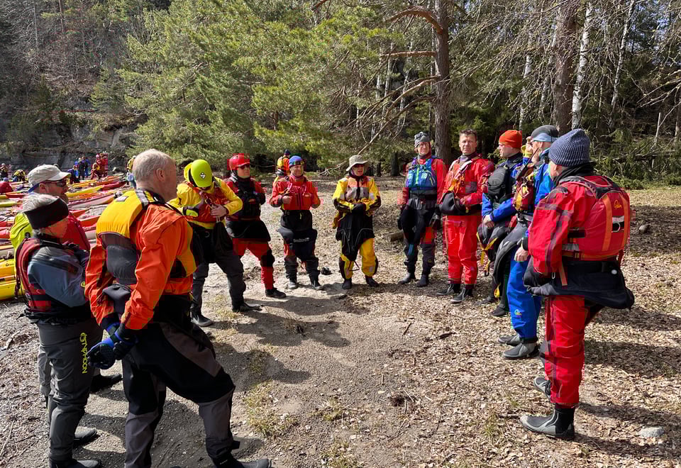 Bildet viser en rekke padlere på land, de står og lytter til en som prater.