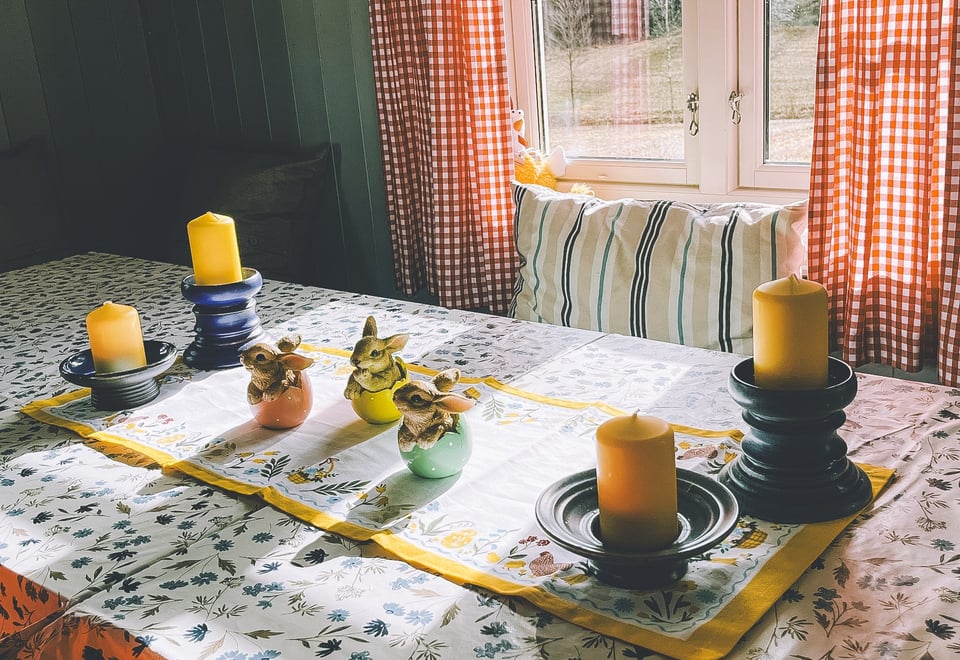 Et bord pyntet med påskeduk, kyllinger og gule stearinlys. I bakgrunnen et vindu med rutede gardiner.