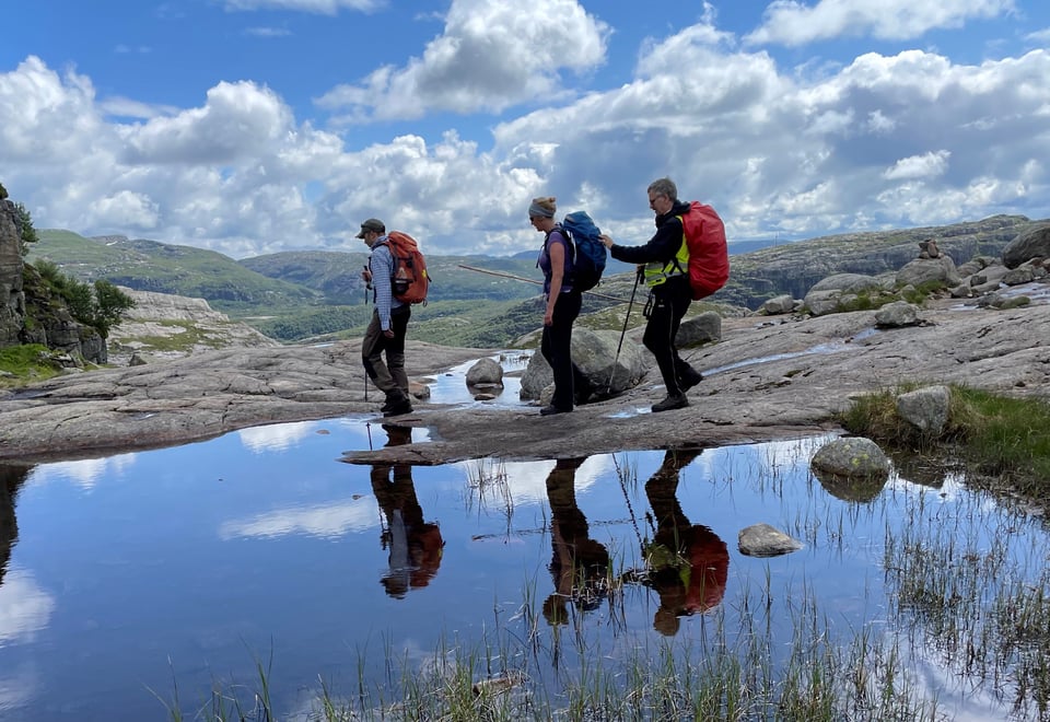 Tre personer går forbi et vann i fjell-landskap. Den bakerste personen på rekka holder i sekken til personen foran og holder en mobilitetsstokk.