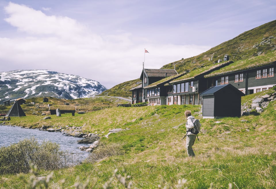 En turgåer med sekk står foran Haukeliseter. Grønt gress og snødekte topper