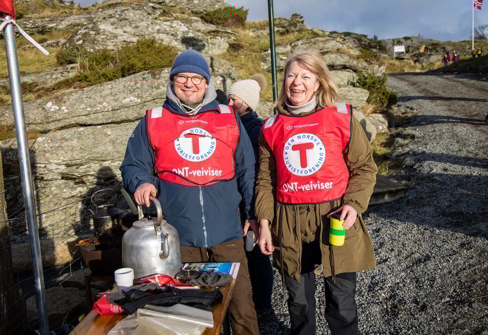 En mann og en kvinne med en rød vest med en stor T på brystet, står og smiler til kamera mens de koker kaffe utendørs.