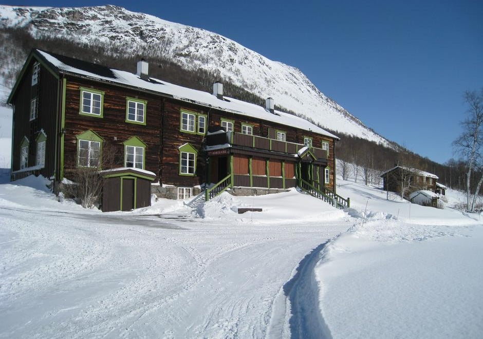 En brun to-etasjes hytte, med to-etasjesveranda foran, med snødekt landskap rundt og et fjell som ruver over bak hytta.