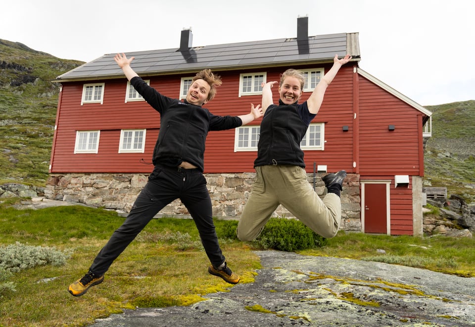 En mann og en dame som hopper med armene i været foran en rødmalt hytte