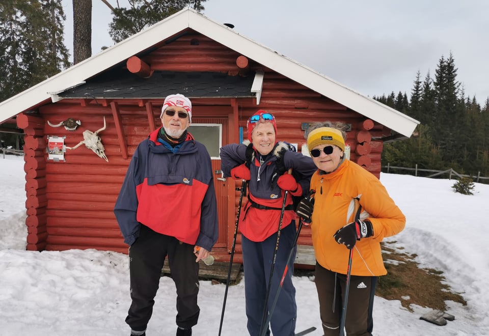 Tre blide eldre mennesker utenfor rød koie.