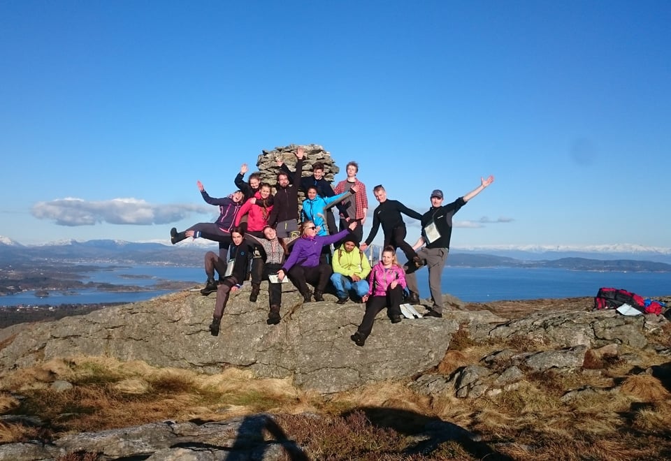 Fjorten ungdommer står foran en varde på en fjelltopp og vinker. Det er sommer, sol, og alle er glade.