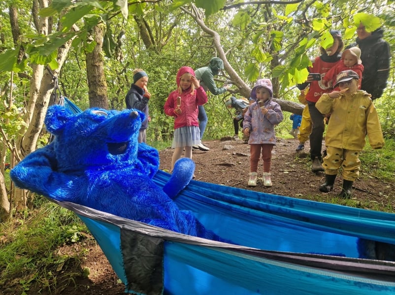 En blå rev slapper av i en hengkøye. Barn spiser is og ser forundret på reven