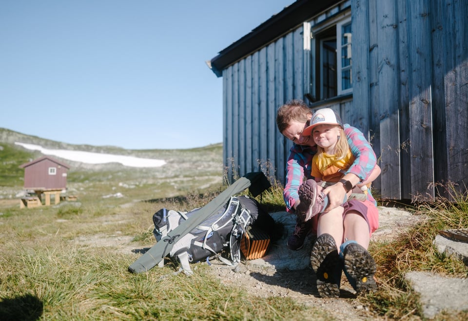 Mor og barn sitter foran hytte i fjellet